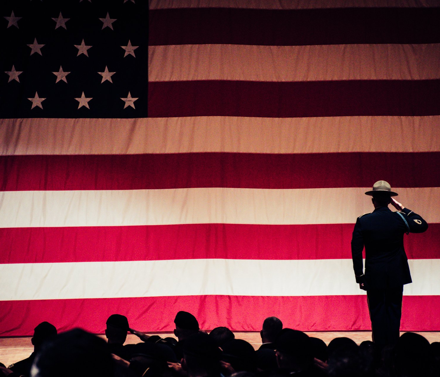 Soldier salutes large American flag; group of people salute in the foreground.