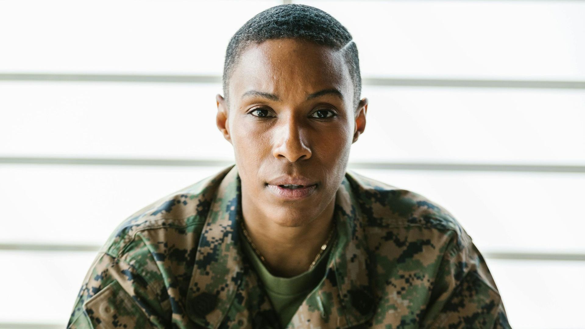 A female soldier is sitting on a stool in front of a window.