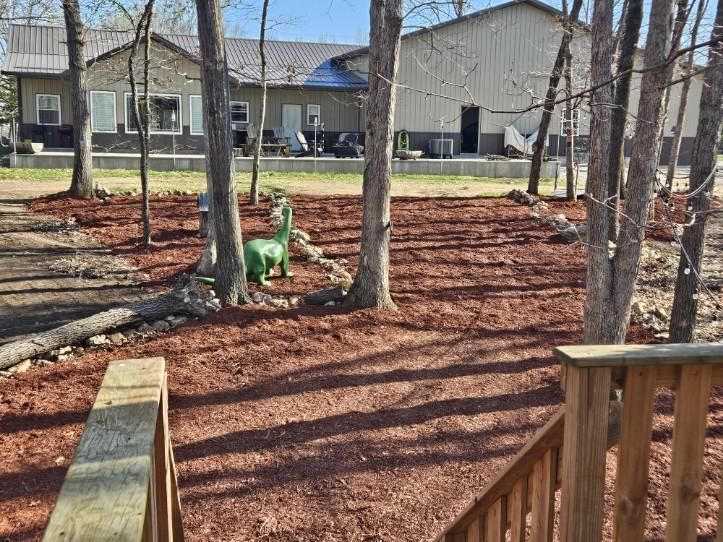 Backyard with mulched ground, trees, and a green dinosaur playground toy near houses