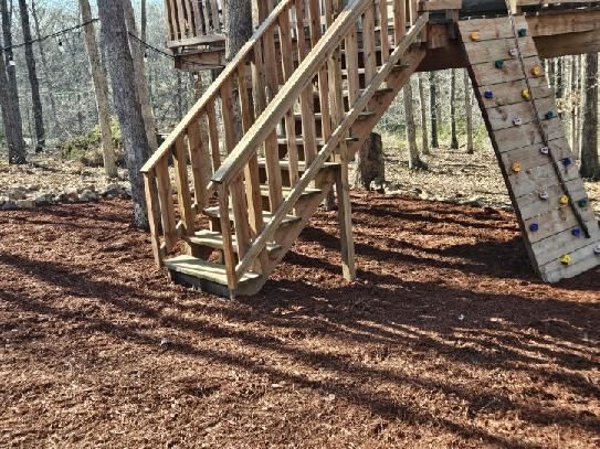 Wooden staircase and climbing wall in a wooded outdoor playground, with leaf-covered ground.