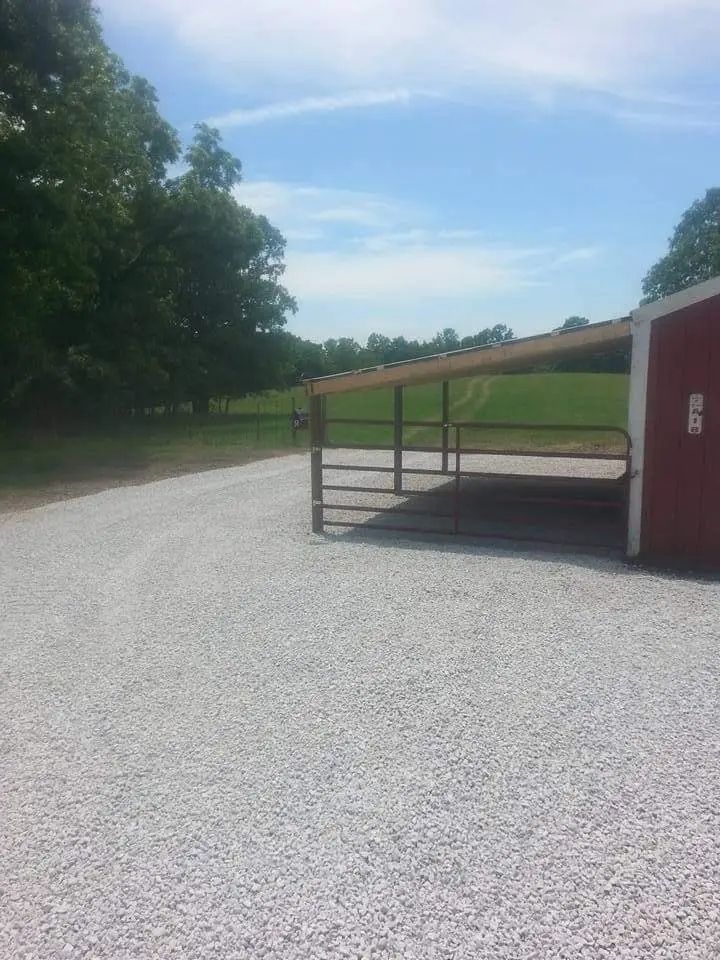 Covered open shelter with metal frame on a gravel lot beside a grassy field and trees.