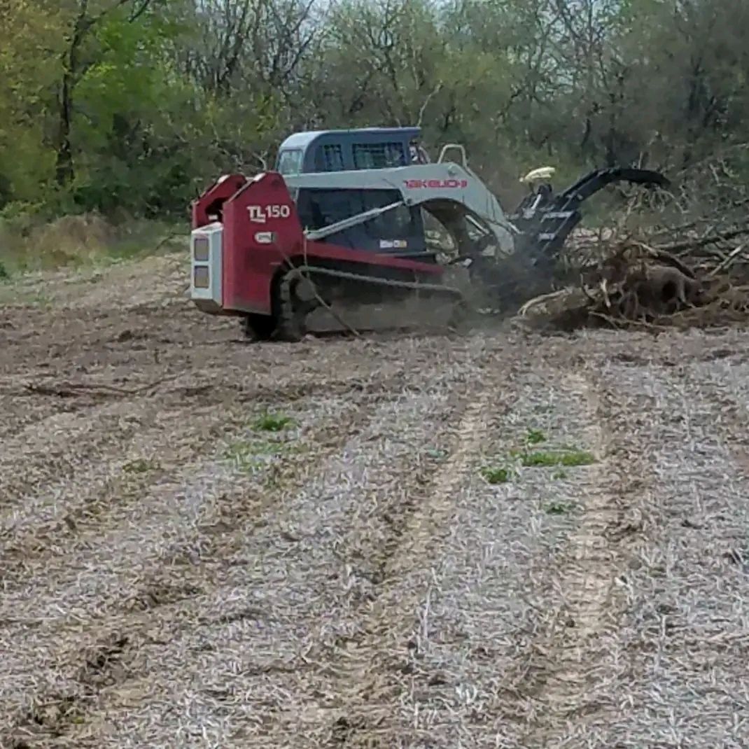 Red excavator clearing brush on a muddy rural path, with a blue truck behind it.