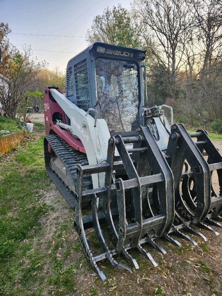 Red skid steer loader with grapple attachment parked on a grassy path by trees