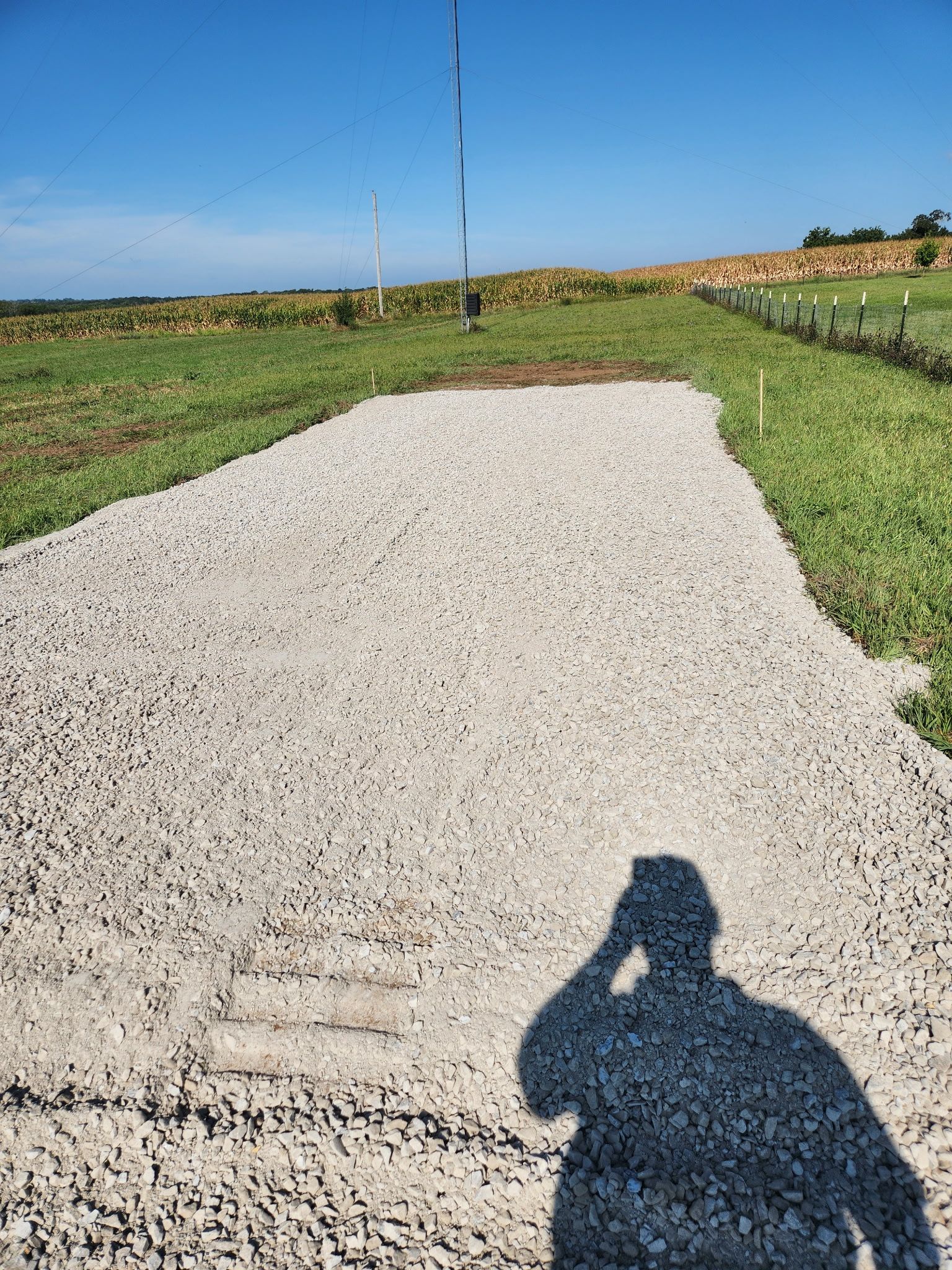 Gravel road leading through green fields under a blue sky, with a person’s shadow in the foreground.
