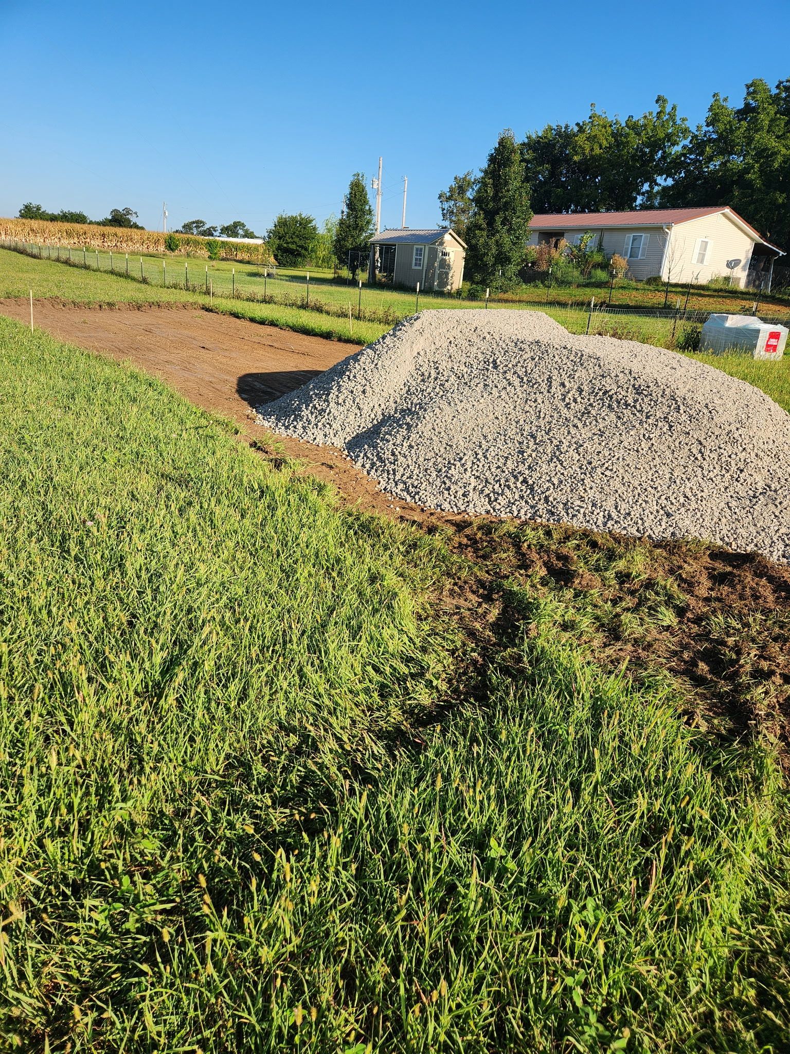 Gravel pile beside a green field under a clear blue sky, with houses and trees in the distance