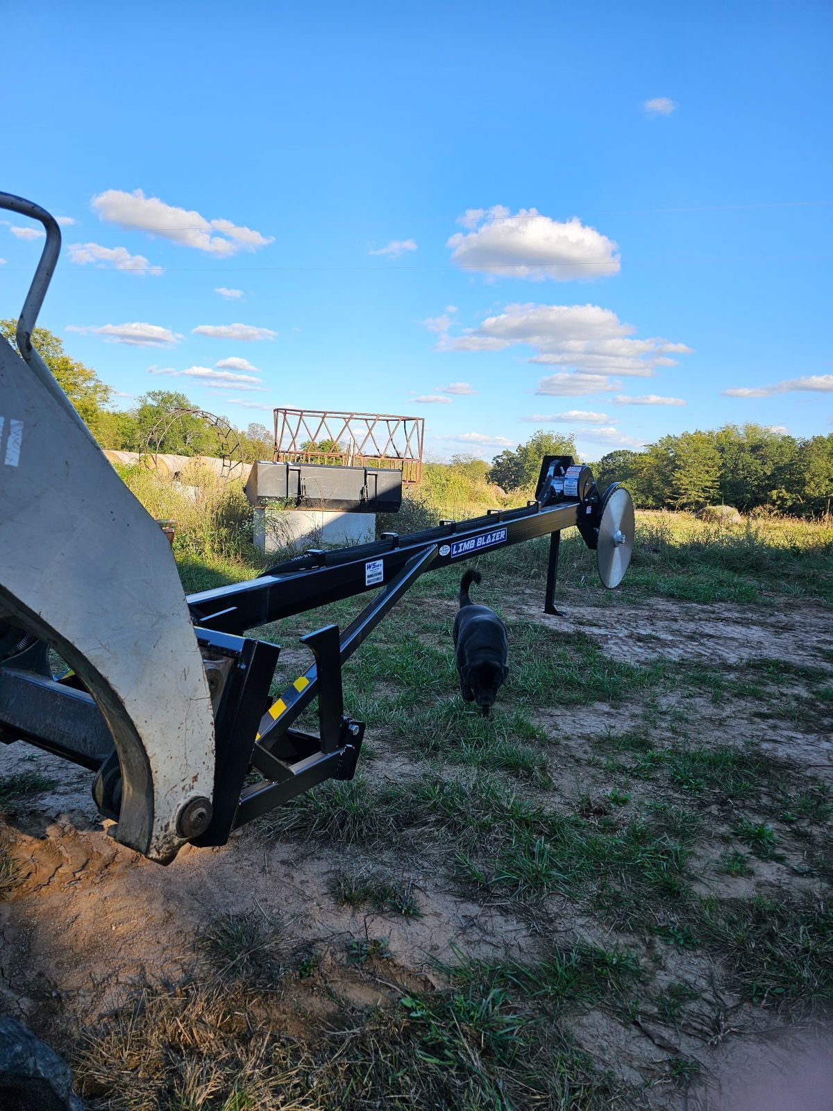 Construction equipment arm in a grassy field under a blue sky, with a person and a dog nearby.