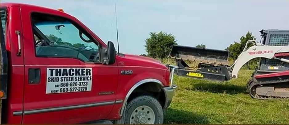 Red Thacker dump truck parked beside a white excavator in a grassy field.