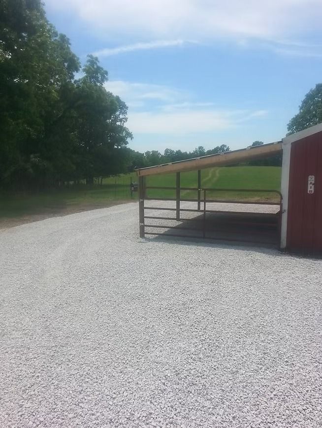 Gravel driveway beside a red barn with a metal gate, trees and blue sky in the background