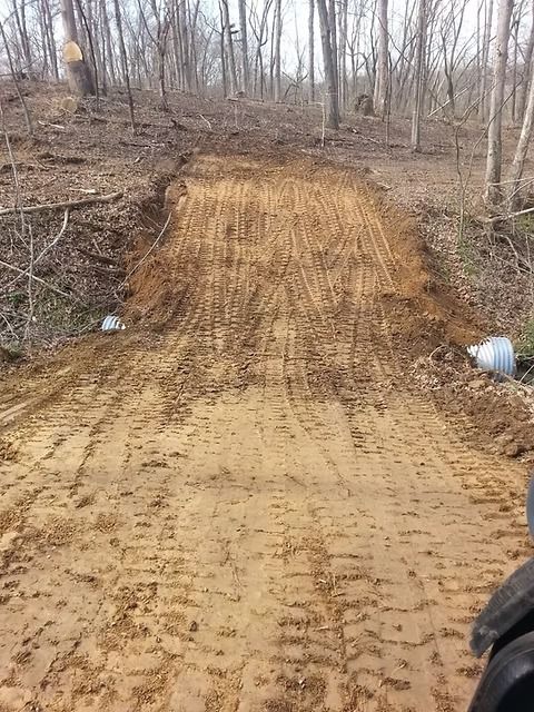 Freshly graded dirt road in a leafless wooded area, with tire tracks and a small white bucket at right.