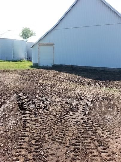 Dirt lot beside a white barn with blue trim and a small door, showing tire tracks in the foreground