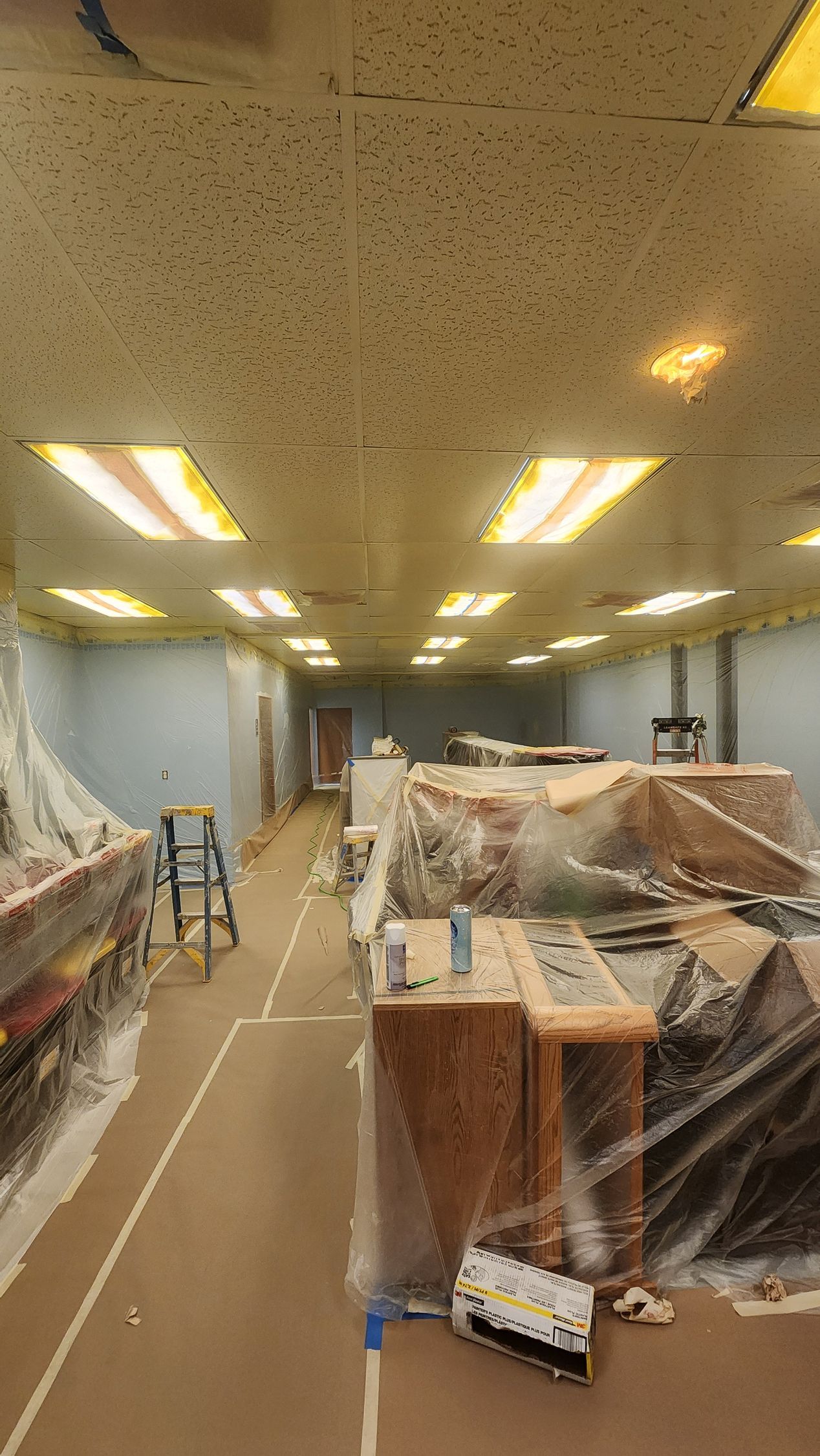 Interior room under renovation, with covered furniture, drop ceiling, and ladders.