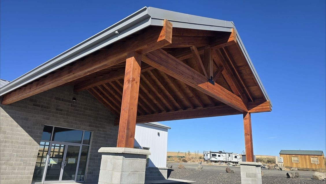 Wooden-framed entrance to a building. Brown beams and rafters with gray roof against a clear blue sky.