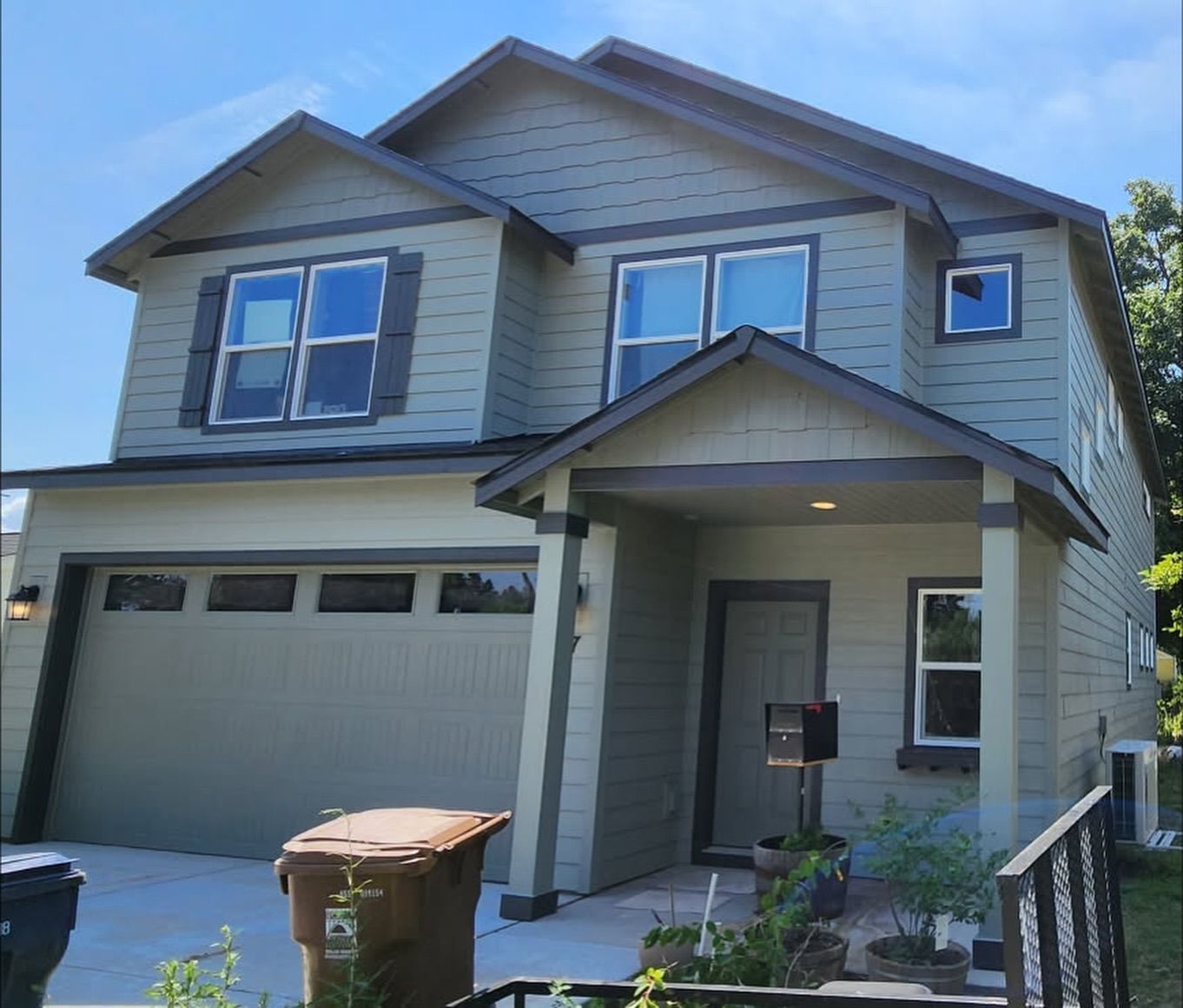 Two-story house with green siding, grey trim, and a brown garage door. A trash can sits in the driveway.
