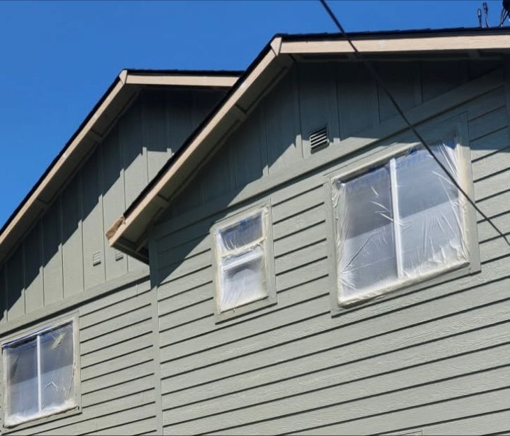 Two-story house with green siding, windows covered in plastic, blue sky.