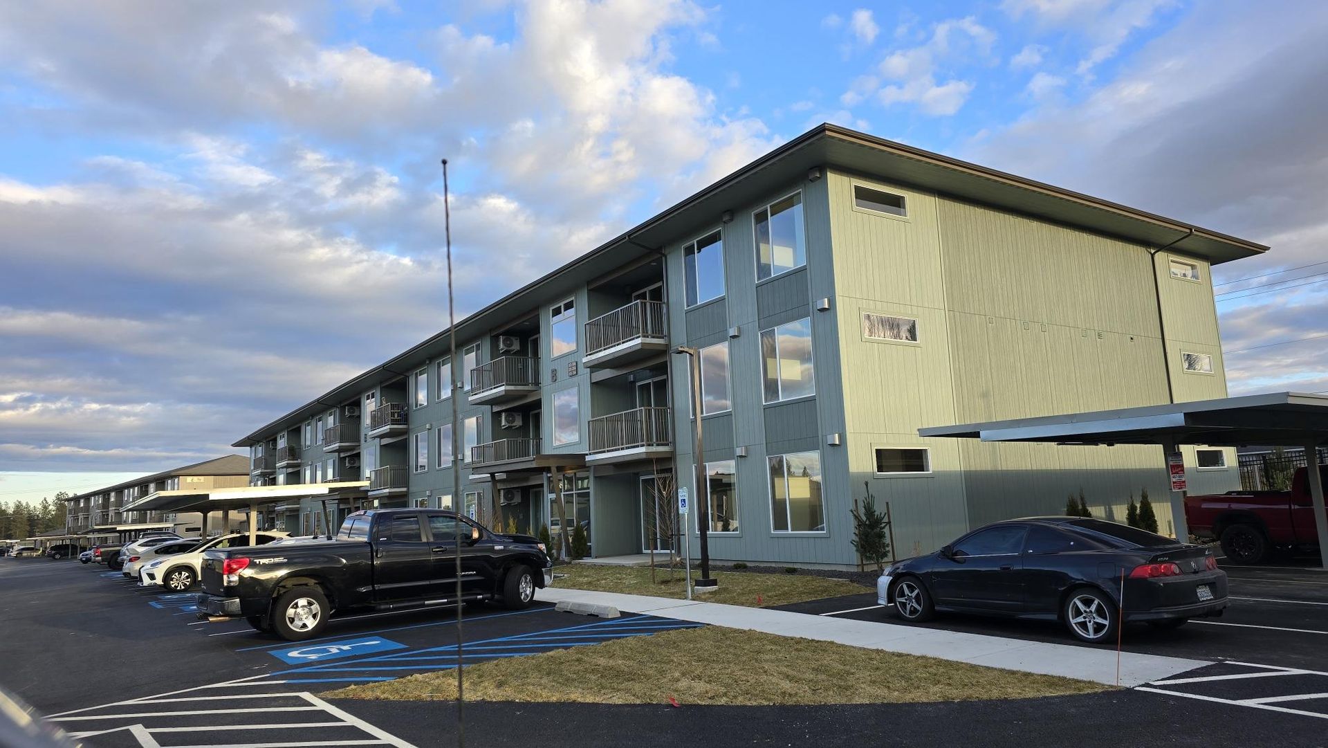 Apartment building with cars parked out front under carports, cloudy sky.
