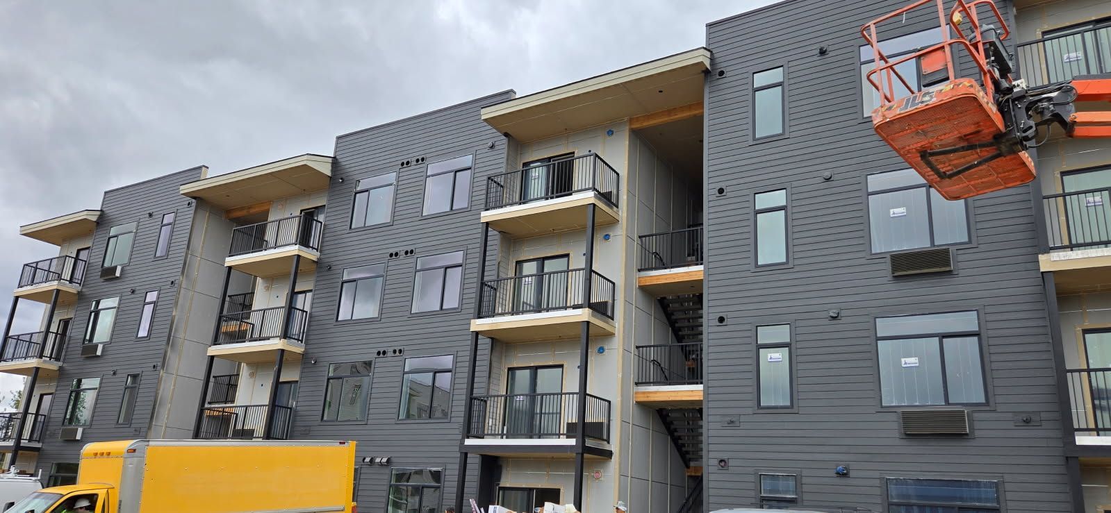 Apartment building under construction; grey siding, multiple balconies, orange lift in the air, yellow truck below.