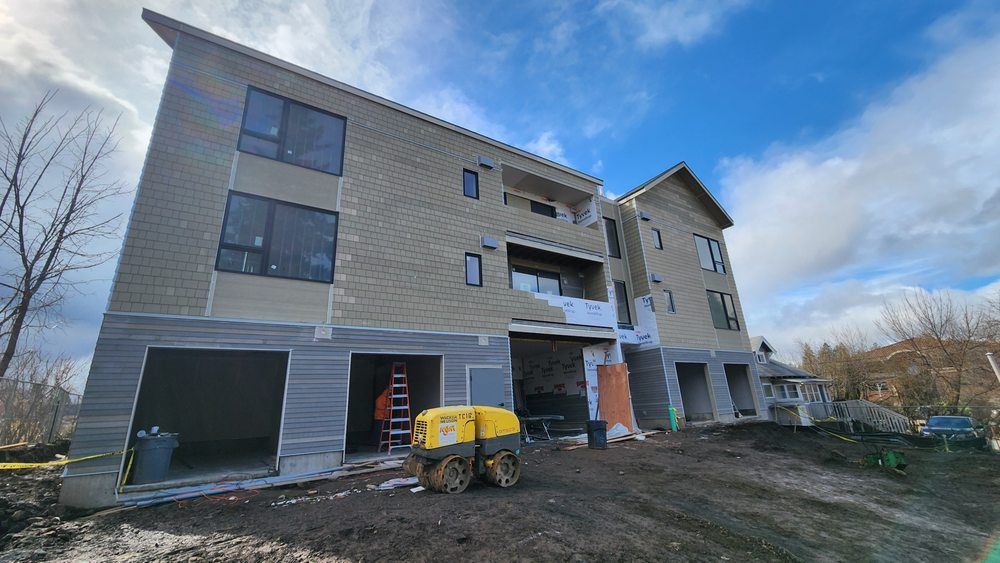 Multi-story building under construction with unfinished stucco exterior, garage bays, and a compact excavator.