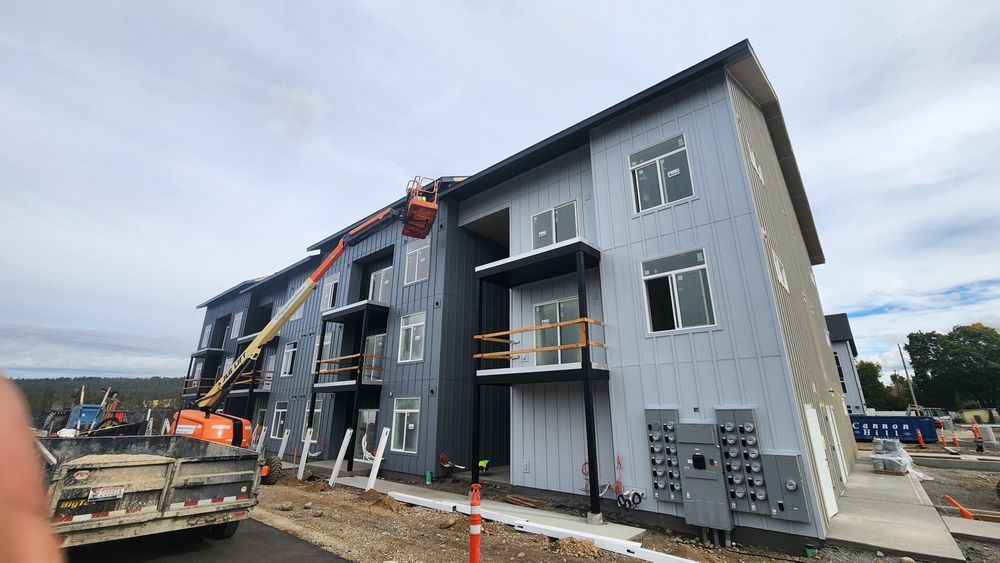 Construction of a multi-story apartment building. A lift carries a worker near the roof, blue-gray siding, overcast sky.
