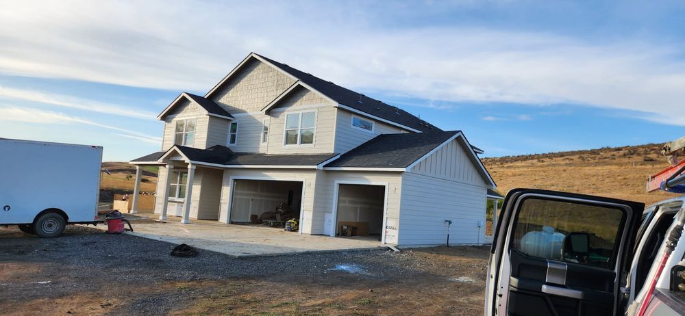Two-story light gray house under construction with attached garage on a gravel lot; white trailer on the left.