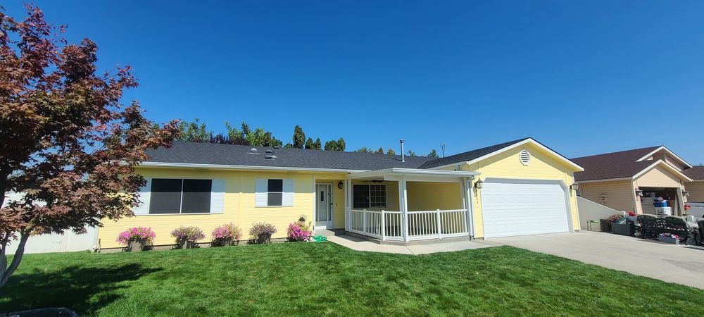 Yellow house with a white garage door, surrounded by green grass and pink flowers under a blue sky.