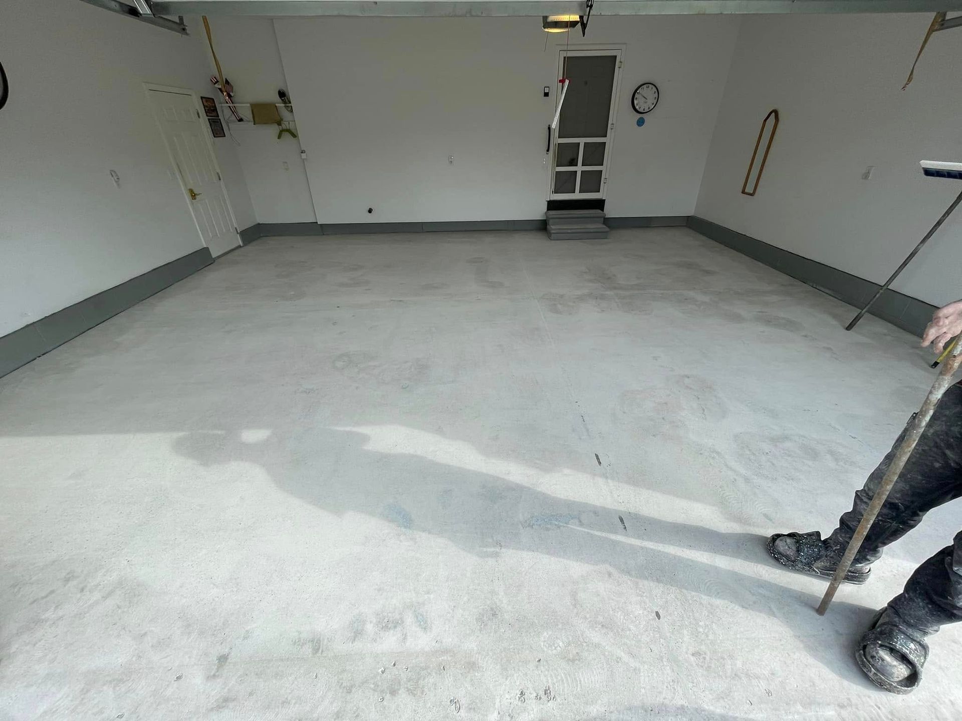 A person in a garage smoothing concrete with a long stick. Concrete floor and white walls.