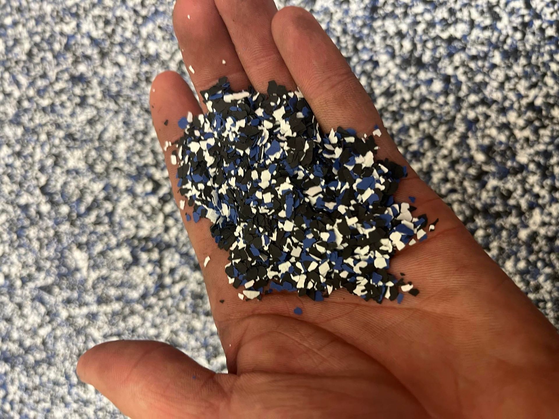 Hand holding a mix of blue, black, and white decorative flakes, possibly for flooring.