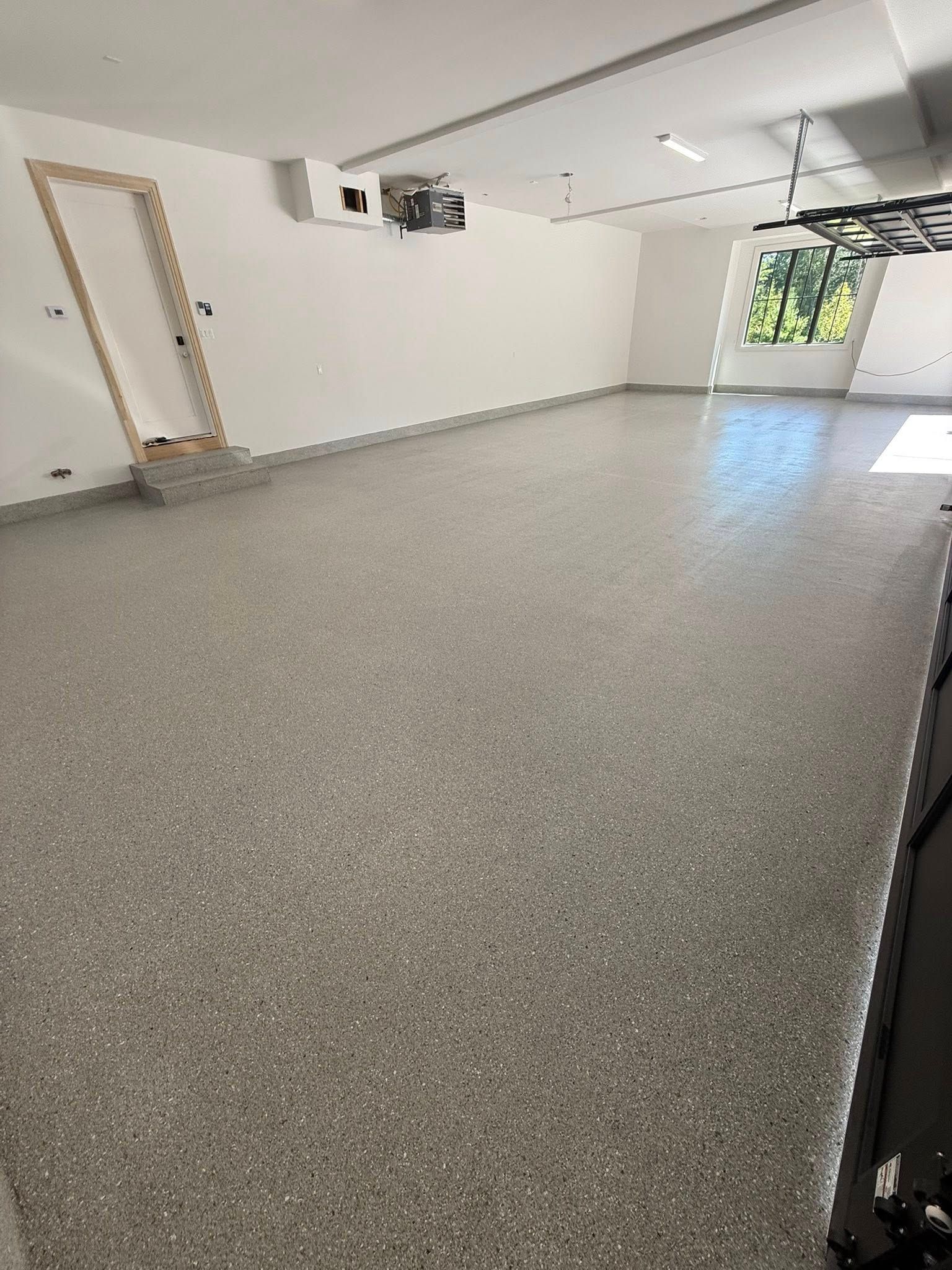 Spacious room with speckled gray flooring, white walls, and a doorway. Sunlight streams in through a window.