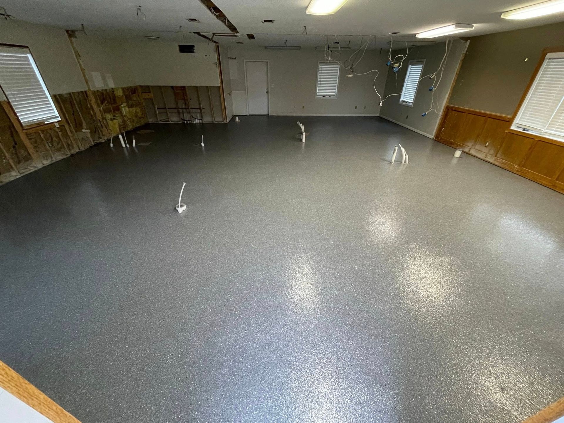 A large room with a gray, speckled epoxy floor, wood paneling, and white-framed windows.