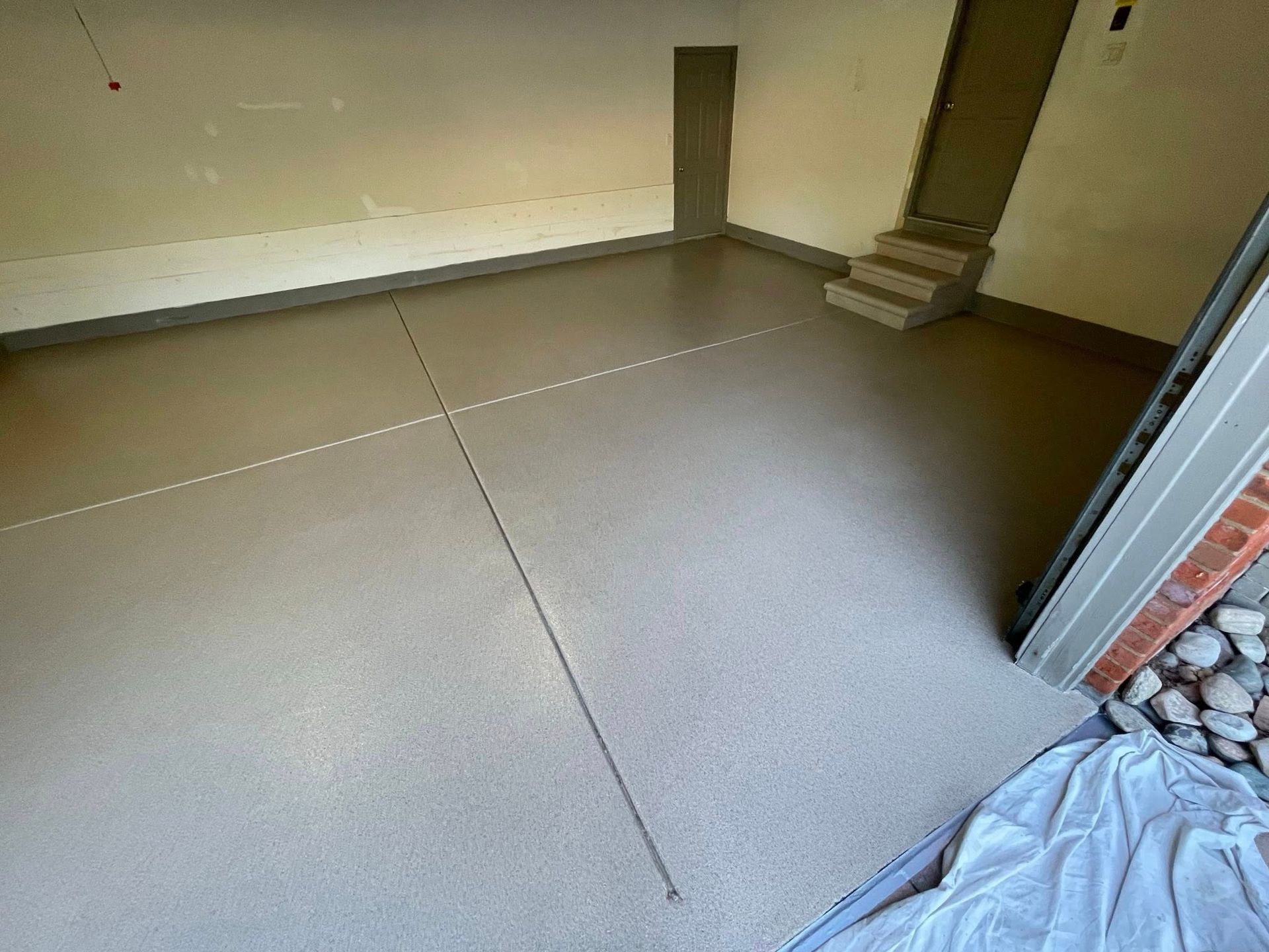 A garage with a fresh, smooth, grey concrete floor. Walls are light tan. A door and steps are visible in the background.