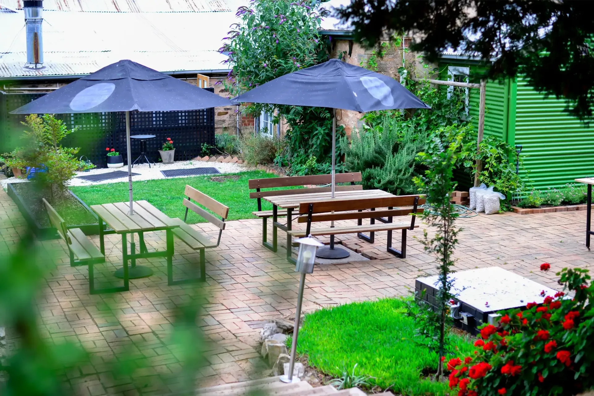 A patio with tables and benches and umbrellas in a garden.
