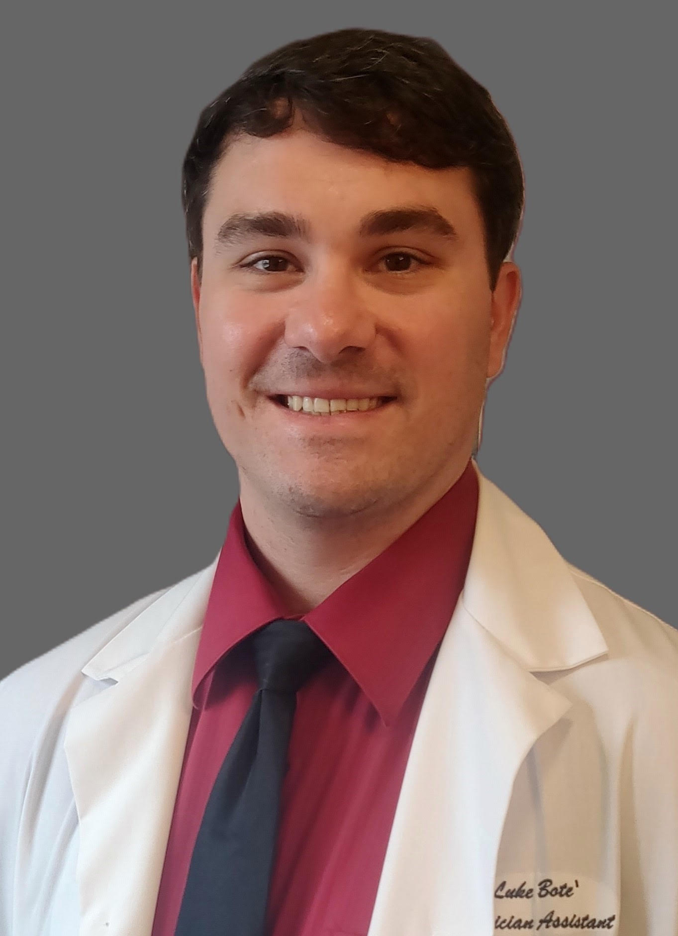 A man, Luke Bote', wearing a lab coat and a red shirt is smiling