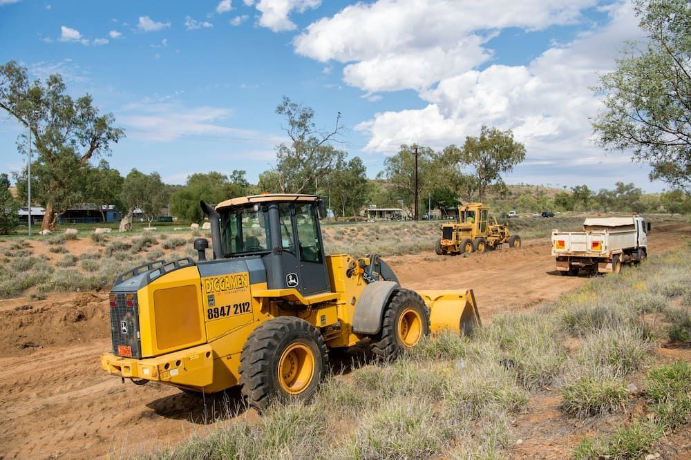 A Yellow Tractor is Driving Down a Dirt Road Next to a Truck — Diggamen Civil Contracting in Tivendale, NT
