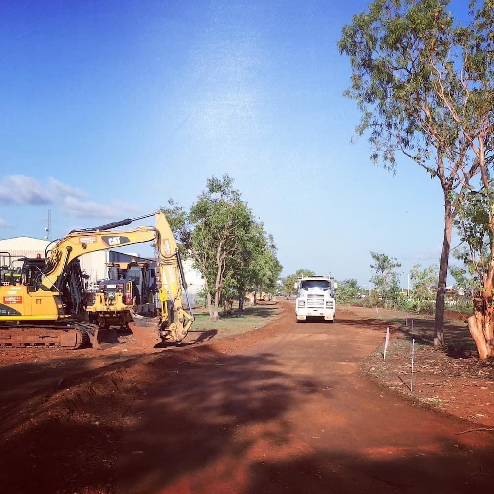 A White Truck is Driving Down a Dirt Road Next to a Yellow Excavator — Diggamen Civil Contracting in Tivendale, NT