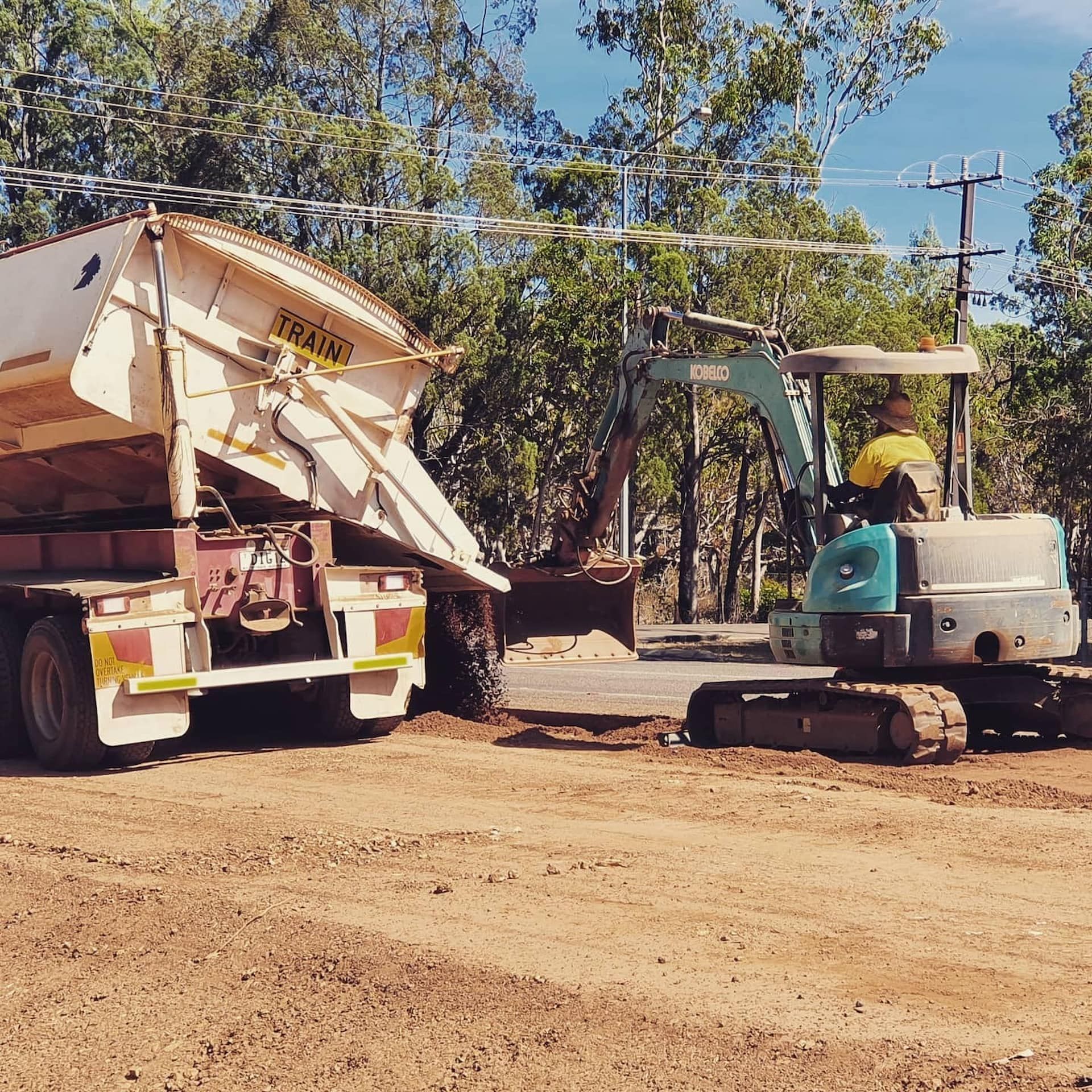 A Man Is Driving an Excavator Next to A Dump Truck — Diggamen Civil Contracting in Tennant Creek, NT