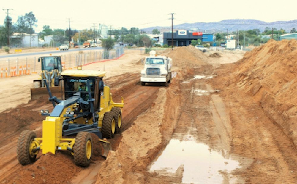 A Yellow Tractor Is Driving Down a Dirt Road Next to A White Truck — Diggamen Civil Contracting in Palmerston, NT