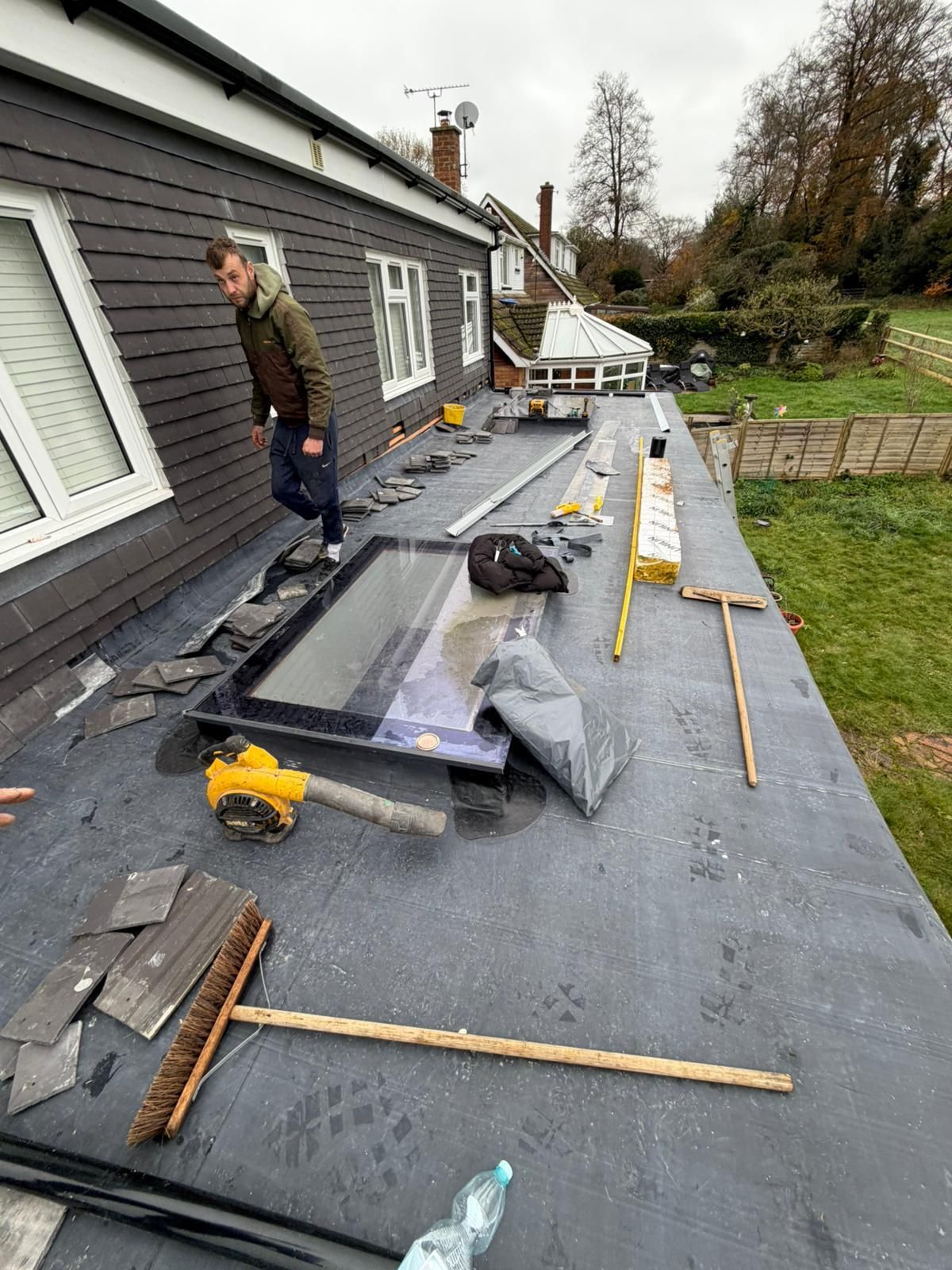 Person on a flat roof with tools and a skylight; a building with dark siding is in view.