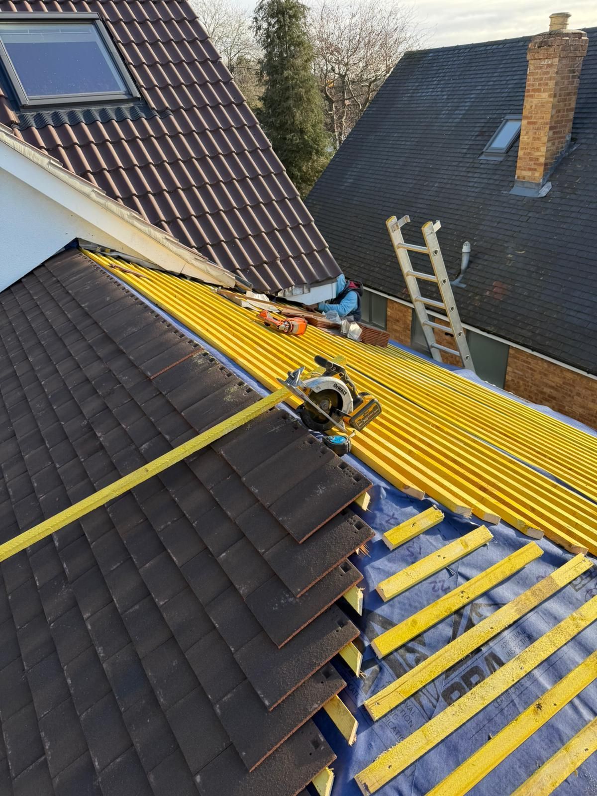Roofers working on a house roof. Yellow corrugated underlayment, tools, and a ladder are visible.