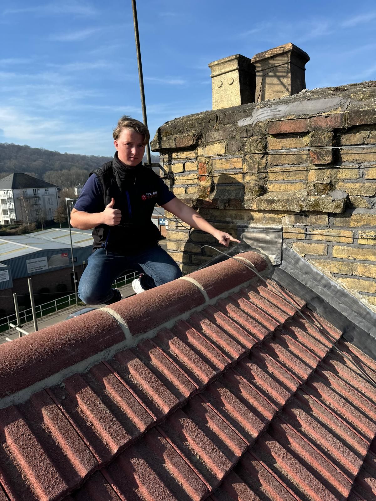 Person kneeling on a tiled roof, pointing; chimney in background.