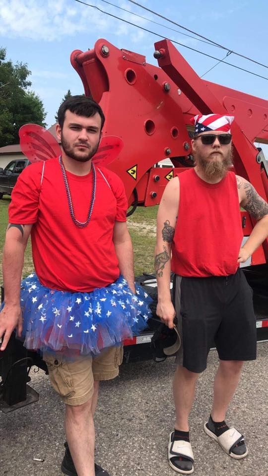 Two men are standing next to each other in front of a tow truck . one of the men is wearing a tutu.