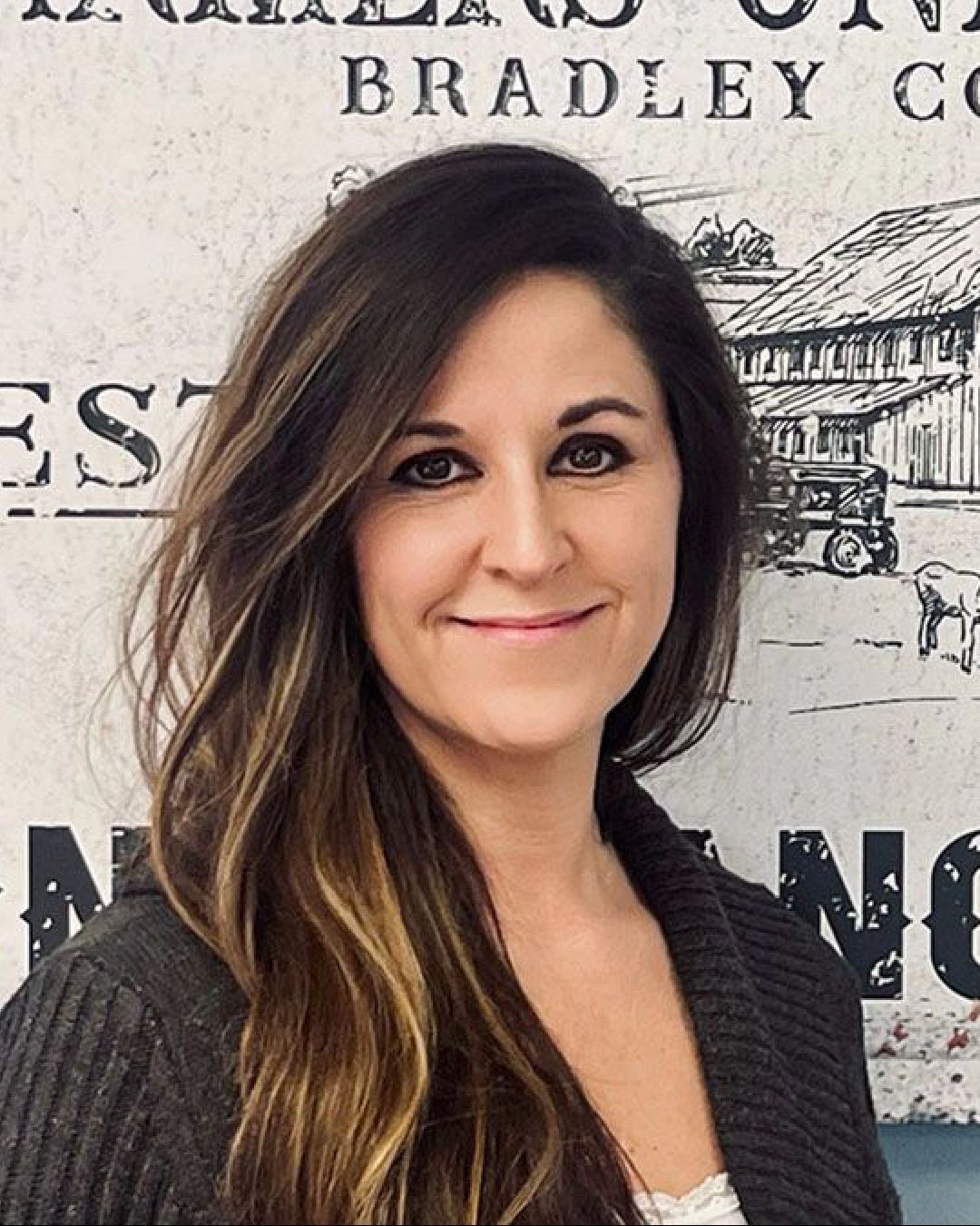A woman with long hair is smiling in front of a wall with a picture of a barn on it.