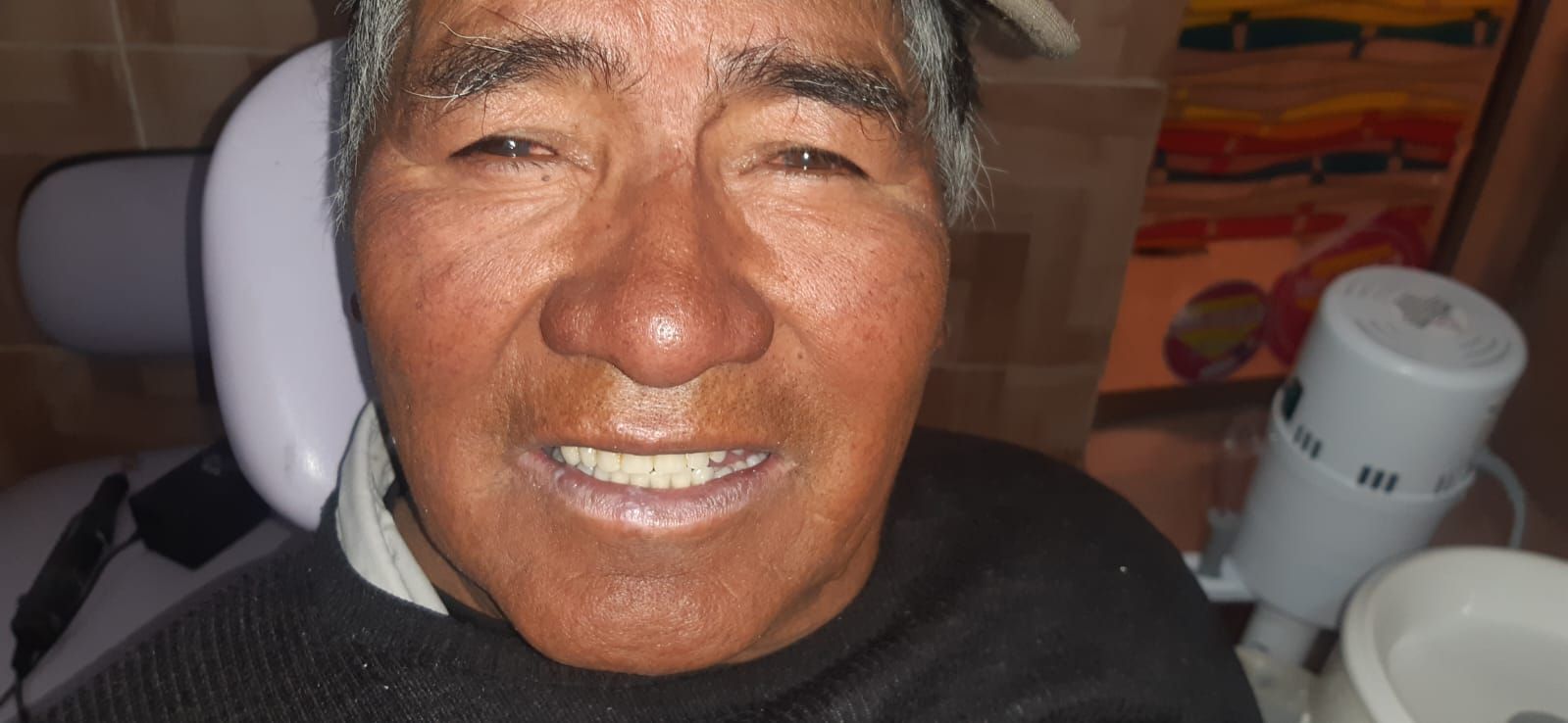 Hombre en un sillón dental, sonriendo, mirando a la cámara. Equipo dental al fondo.