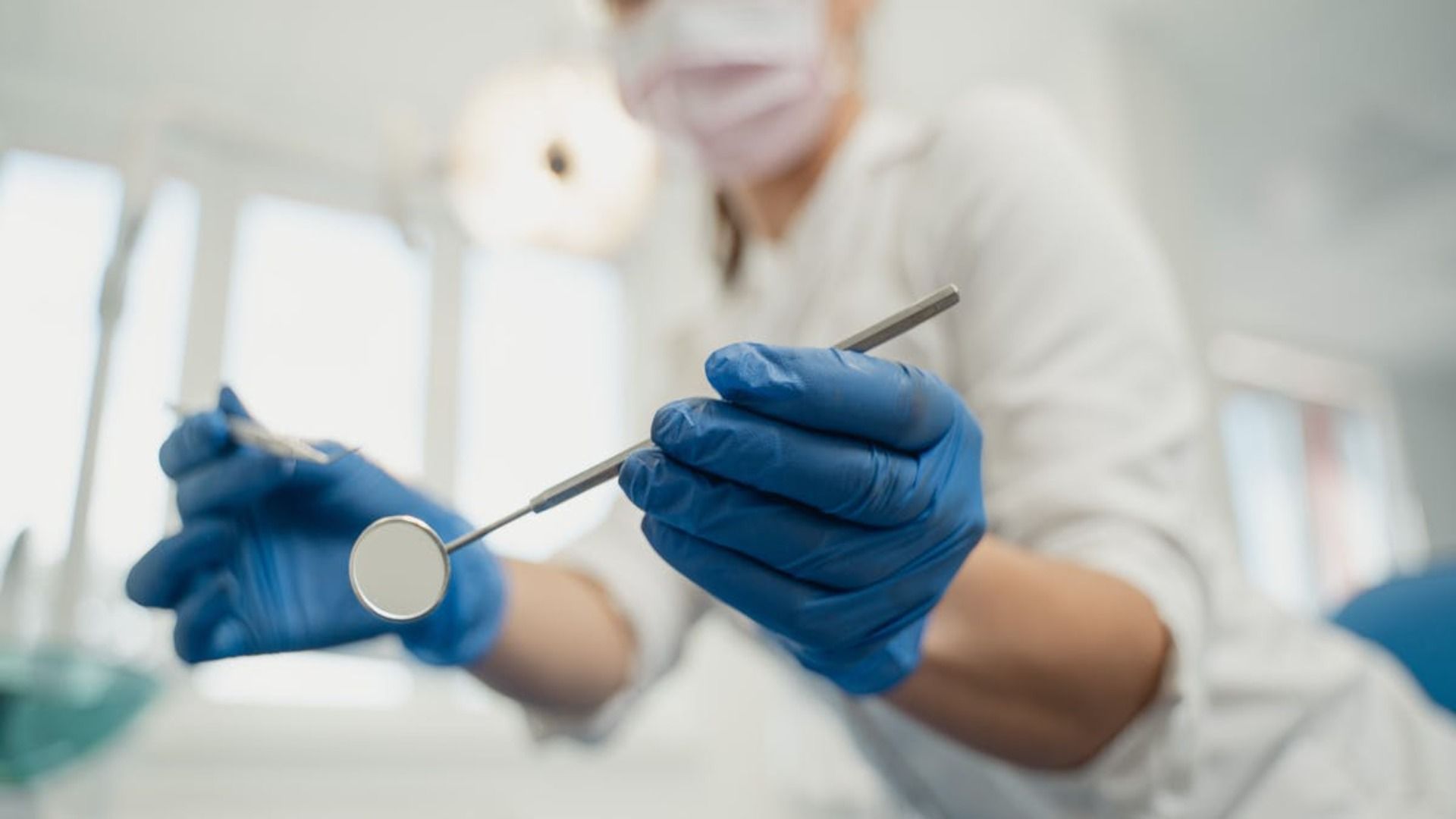 Dentista con bata blanca y mascarilla sostiene espejo dental, guantes azules, interior luminoso.