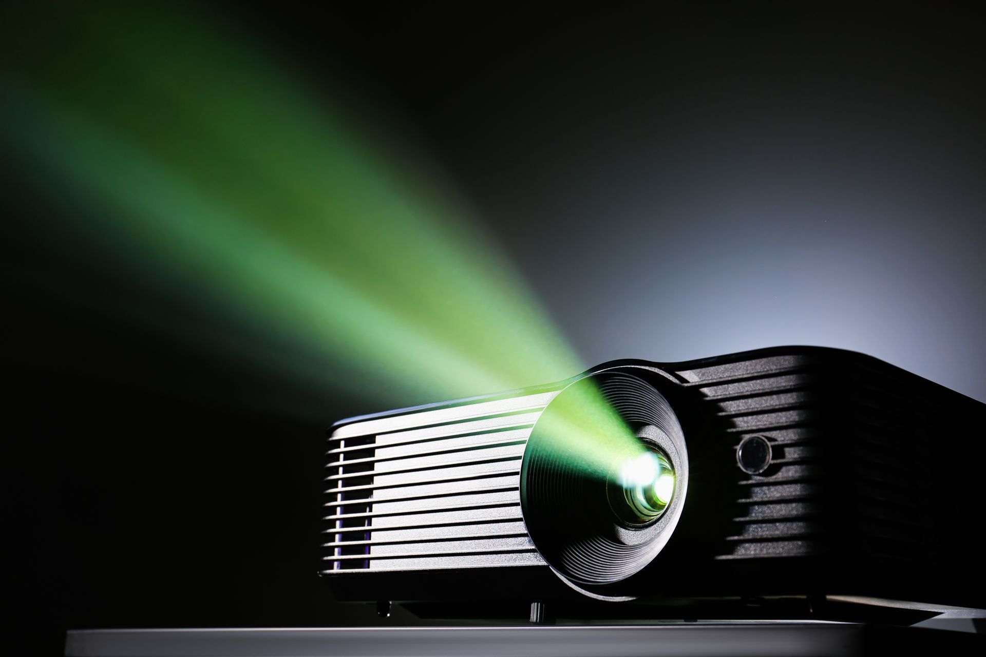 A black digital projector sits on a flat surface, casting a bright, luminous green light beam into the dark room.