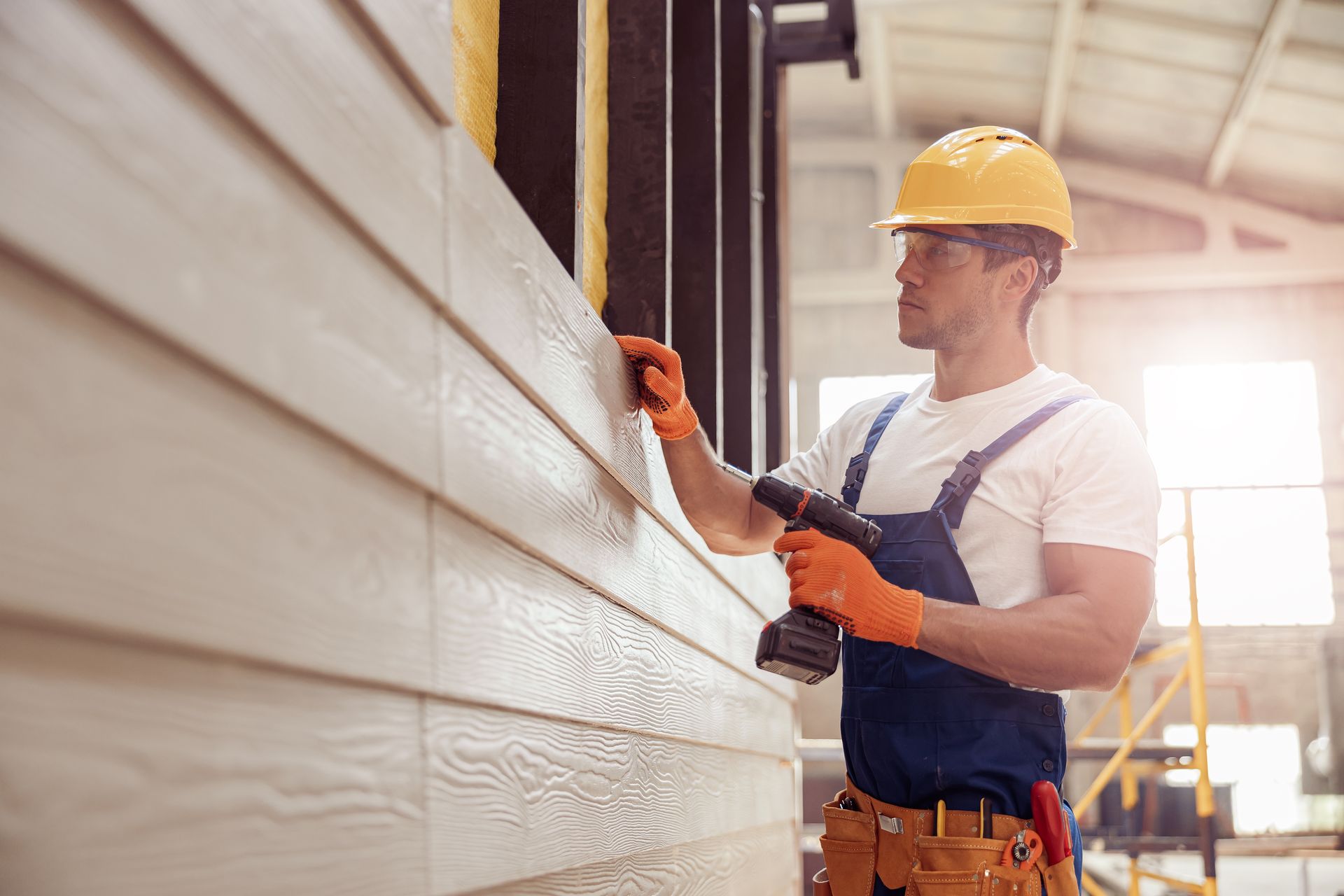 Construction worker installing siding with a drill. Wearing a yellow hard hat and orange gloves.