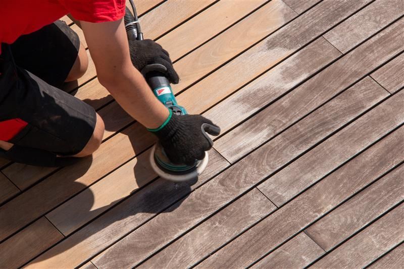 Person sanding wooden deck with electric sander, wearing black gloves.