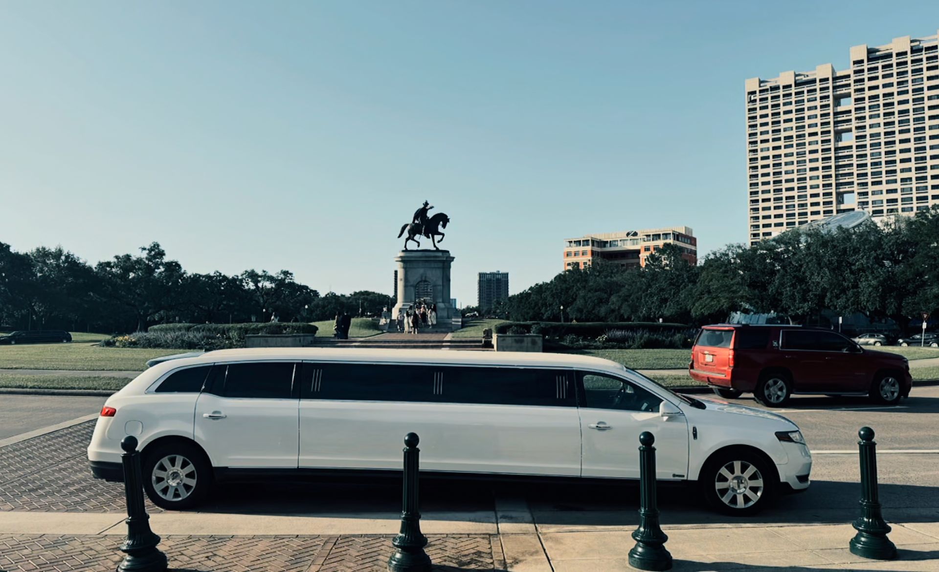 White limousine parked, equestrian statue and buildings in background, sunny day.