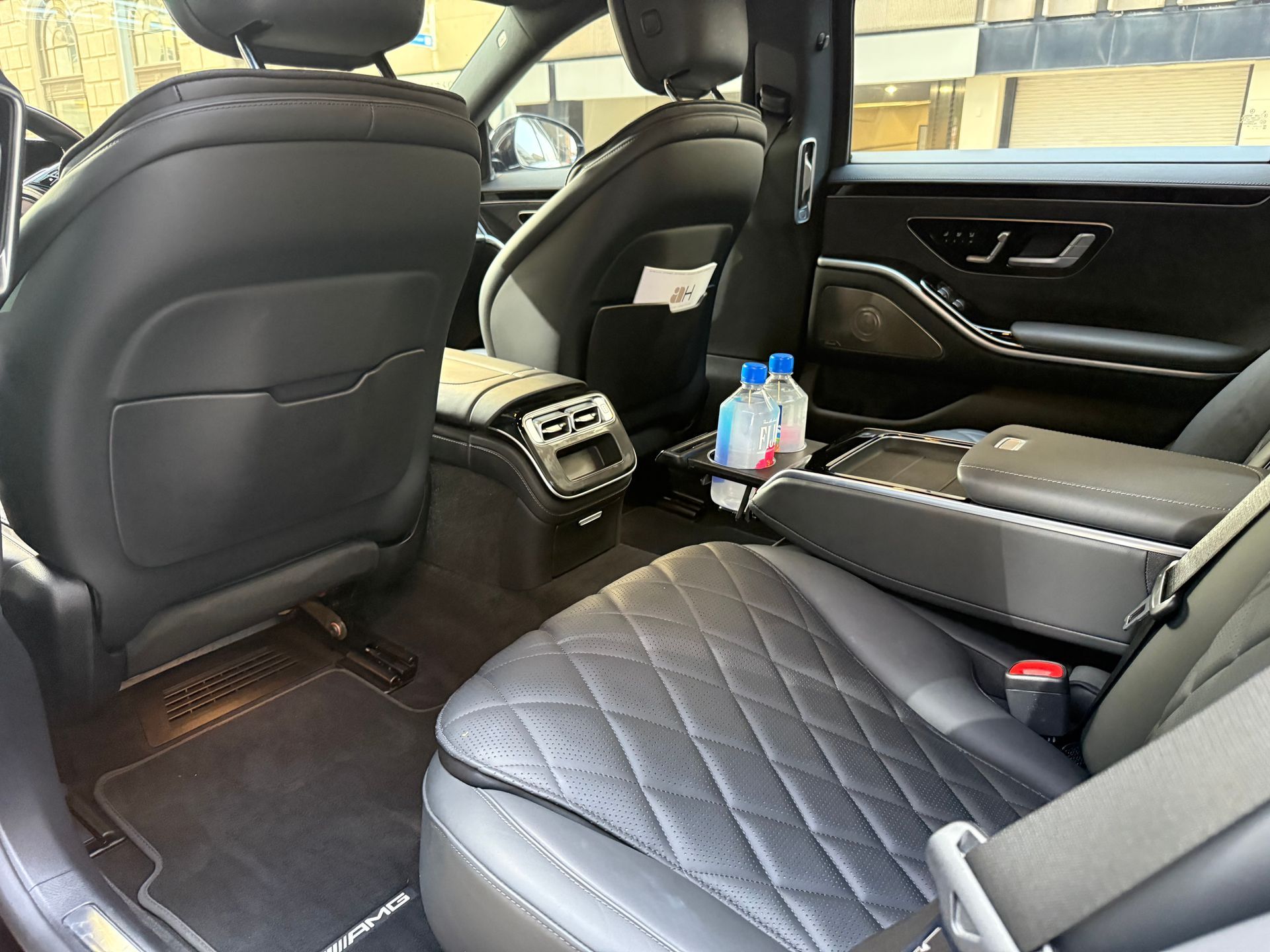 Rear interior of a black Mercedes-Benz S-Class with quilted leather seats, a center console, and water bottles.