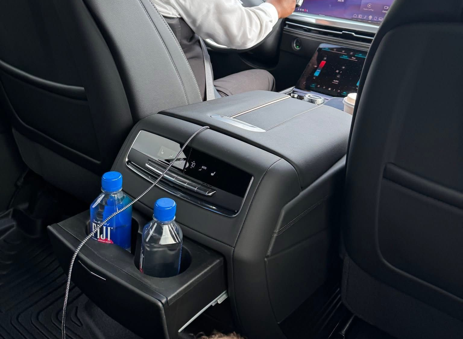 Rear interior of a vehicle with a center console holding water bottles and a charging cable. Person in the front.