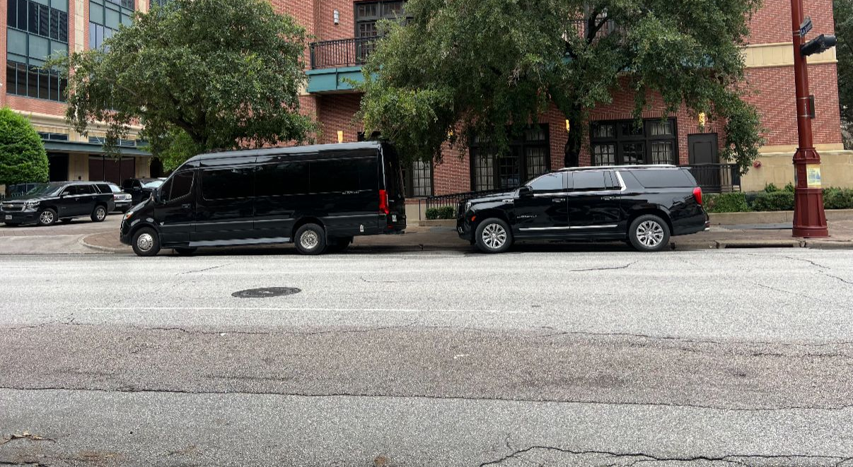 Black van and SUV parked on a city street with a brick building and trees in the background.