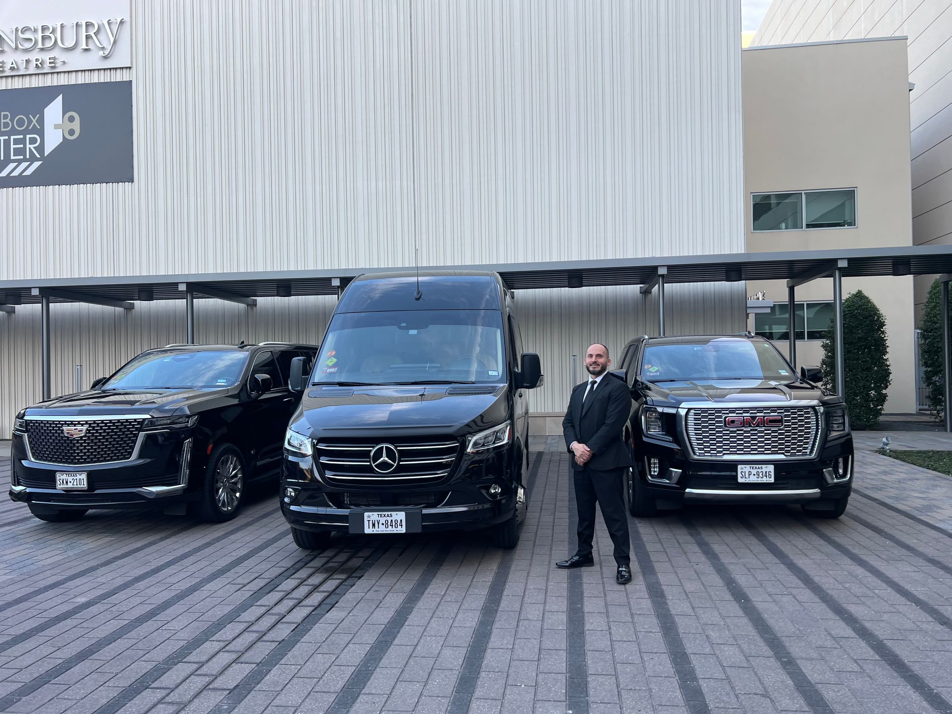 A man stands between three black vehicles outside a building.